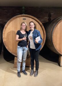 Céline en Ambra staan met een glas wijn in de hand in de wijnkelder voor een groot houten wijnvat. 